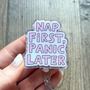 May include: A badge reel featuring a glittery, translucent acrylic topper with the text NAP FIRST, PANIC LATER in light purple, sans-serif capital letters. The badge reel includes a clear plastic strap and a metal clip for attachment.
