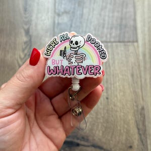 May include: A retractable badge reel with a skeleton design. The badge reel features a skeleton giving a thumbs up, a rainbow, and the text "WE'RE ALL DOOMED BUT WHATEVER" in pink and white. The reel is held by a hand.