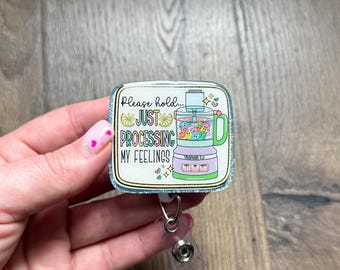May include: A white and green glitter badge holder with a food processor illustration and the text "Please hold... Just processing my feelings".