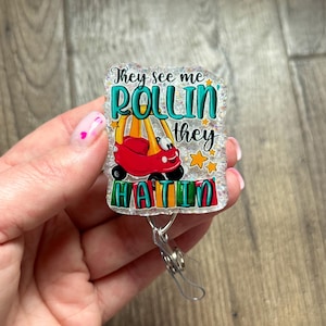 May include: A white glitter badge holder with a red toy car and the text "They see me rollin' they hatin'".