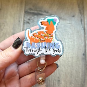 May include: A badge reel featuring a cartoon dachshund wearing a winter hat and a string of lights. The text "Dashing through the snow" is written in blue and white. The reel has a clear plastic holder and a gold-colored metal clip.
