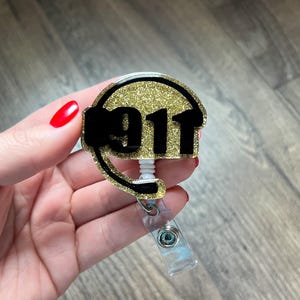 May include: A gold glitter badge reel with the number "911" in black. The badge reel has a black phone receiver design around the numbers and a clear plastic retractable cord and clip.