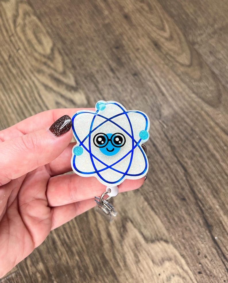 May include: A white glitter atom with a blue outline and a blue smiling face with glasses. The atom is attached to a white retractable badge holder.