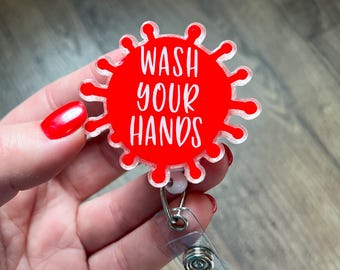 May include: A red badge reel with the words "WASH YOUR HANDS" in white. The badge is shaped like a virus and has a clear plastic clip and retractable cord.