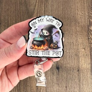 May include: A black and white Halloween-themed retractable badge holder with a cartoon Grim Reaper stirring a cauldron. The text on the badge holder reads "On my way to stir the pot."