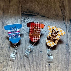 May include: Three badge reels in the shape of baby onesies. One is blue with pink stripes and the text "I ♡ Tiny Humans". The second is red plaid with the same text. The third is yellow and white checkered with the same text.