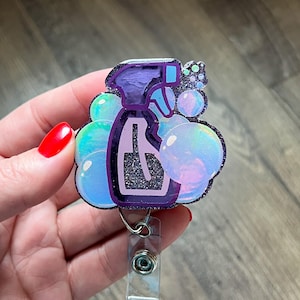 May include: A badge reel featuring a purple spray bottle design with iridescent bubbles. The badge reel has a silver clip and a clear plastic holder for an ID badge. The spray bottle has a glittery black outline.