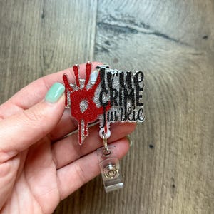 May include: A silver and black glitter badge reel with a red handprint and the text "True Crime Junkie".