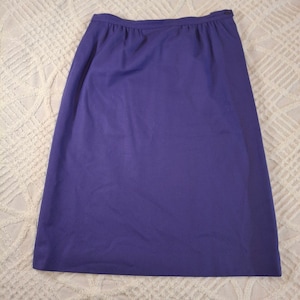May include: A solid purple knee-length skirt featuring a high waistband with subtle gathering at the top. The skirt has a straight silhouette and a simple, clean design with a small button closure visible on the right side of the waistband.