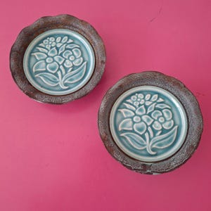 May include: Two circular coasters featuring a central light blue glazed ceramic inlay with a raised floral relief pattern. The outer rim is made of a dark, textured, rust-colored material with a scalloped edge, providing a contrast.