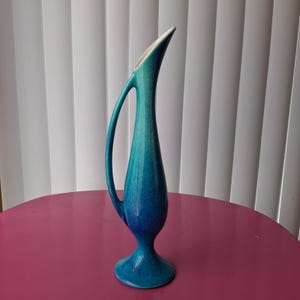May include: A tall, slender ceramic vase featuring a vibrant teal and blue gradient glaze. The vase has a distinctive elongated neck, a single curved handle extending from the body to the upper neck, and a flared base for stability.
