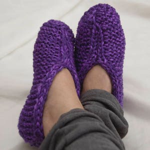 May include: Purple knitted slippers with a cable knit pattern. The slippers are worn on a person's feet.
