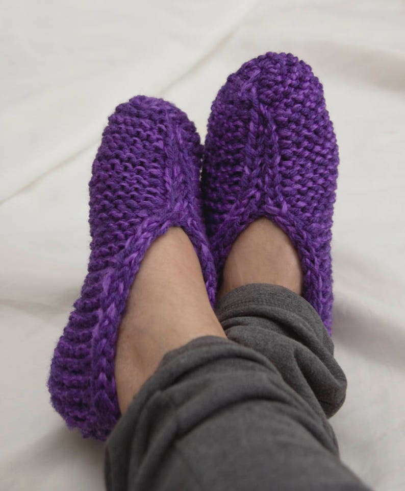 May include: Purple knitted slippers with a cable knit pattern. The slippers are worn on a person's feet.