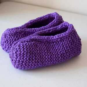 May include: A pair of purple knitted slippers. The slippers are made with a simple knit stitch and have a soft, cozy feel.