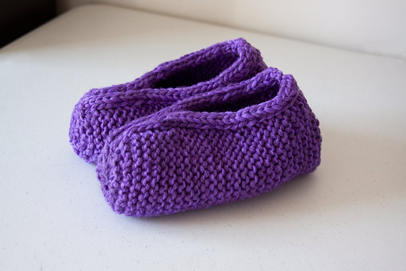 May include: A pair of purple knitted slippers. The slippers are made with a simple knit stitch and have a soft, cozy feel.