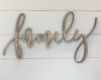 May include: Wooden 'family' sign in a cursive font. The sign is made of light-coloured wood and is mounted on a white, horizontal-planked background. The word 'family' is the central focus, ideal for home decor.