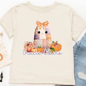 May include: Cream-colored t-shirt featuring a patchwork ghost design with a bow, pumpkins, and floral accents. The text "Gracie Claire" is below the graphic. A "Hey Boo" banner and denim jeans are also visible.