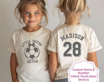 May include: Two white t-shirts with a soccer theme. One shirt says "Little Sister Big Fan" with a soccer ball and a bow. The other shirt says "Madison 28".