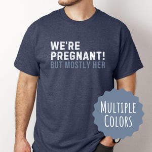 Peut inclure: Un t-shirt bleu avec le texte "We're Pregnant! But Mostly Her" en blanc. Le texte a un aspect vieilli et délabré. Le t-shirt est disponible en plusieurs couleurs.