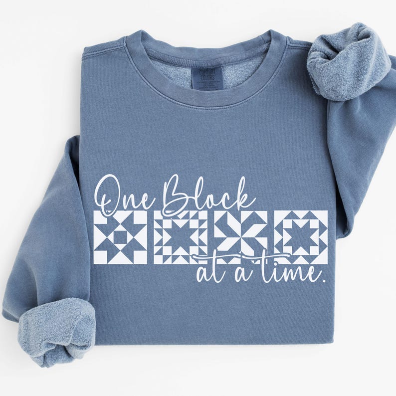 May include: A blue sweatshirt with the text "One Block at a Time" in white script. Below the text are three white quilt block designs. The sleeves are cuffed, and the sweatshirt is on a white surface.