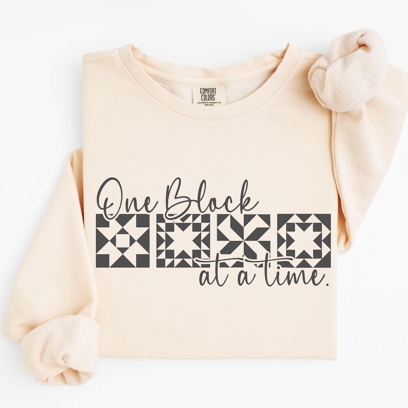May include: A cream-colored sweatshirt with the text "One Block at a time." The text is in a dark gray color and features four quilt block designs. The sweatshirt is folded, with the sleeves visible.