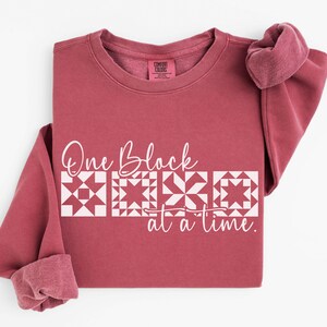 May include: A maroon Comfort Colors sweatshirt with the text "One Block at a time" in white. The text is above a row of four white quilt block designs. The sleeves are cuffed, and the collar tag is visible.