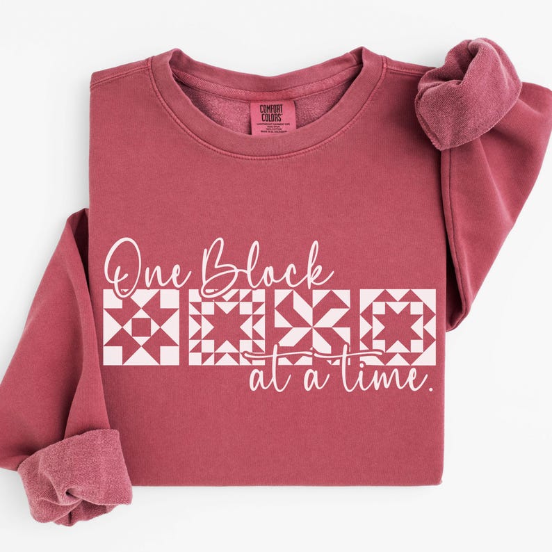 May include: A maroon Comfort Colors sweatshirt with the text "One Block at a time" in white. The text is above a row of four white quilt block designs. The sleeves are cuffed, and the collar tag is visible.