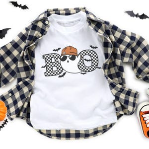 May include: White t-shirt with a "BOO" graphic featuring a ghost wearing sunglasses and a brown cap. The letters are checkered. A blue and tan plaid shirt is layered over the t-shirt. Black bat cutouts are scattered around the shirt, with an orange pumpkin and orange sneakers.