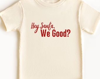 May include: A cream-coloured t-shirt featuring the red text "Hey Santa, We Good?" across the front. The shirt has short sleeves and a crew neck. A festive holiday-themed tee.