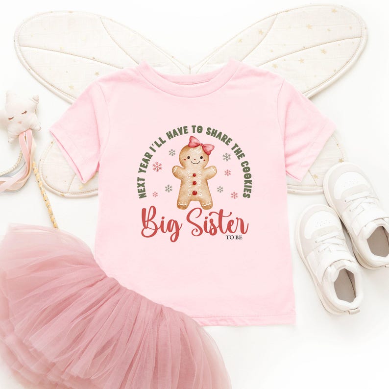 May include: A light pink t-shirt with the text "Next year I'll have to share the cookies" and "Big Sister to be". The shirt features a gingerbread person graphic with a pink bow. The shirt is paired with a pink tutu and white sneakers.