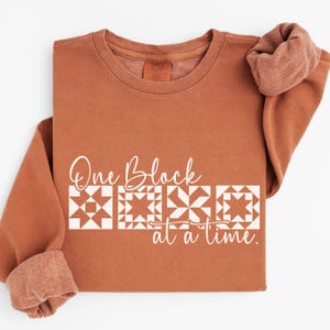 May include: A rust-colored sweatshirt with the text "One Block at a time" in white script. Below the text are three white quilt block designs. The sleeves are rolled up, revealing a soft, textured interior.