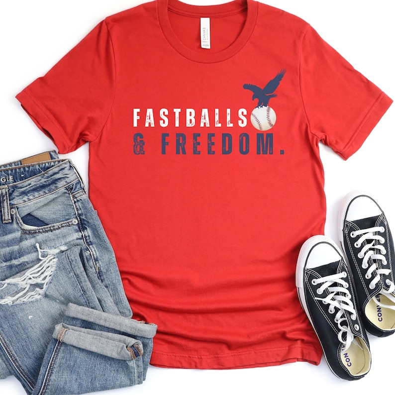 Może przedstawiać: Czerwona koszulka z przetartym niebieskim nadrukiem, na kt&oacute;rym widnieje napis "Fastballs & Freedom." Orzeł leci nad piłką baseballową. Koszulka leży na białym tle, obok pary czarnych but&oacute;w sportowych i niebieskich dżins&oacute;w.
