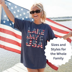 May include: Navy blue t-shirt with the text "LAKE DAY in the USA" in a red, white, and blue design. The person is holding an American flag. The shirt is a casual style.