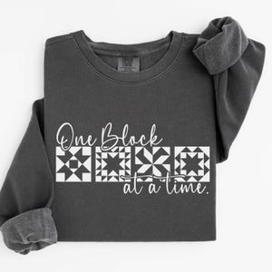 May include: Dark gray sweatshirt with the text "One Block at a Time" in white script and block letters. The design includes four white quilt block patterns. The sweatshirt has a crew neck and long sleeves.