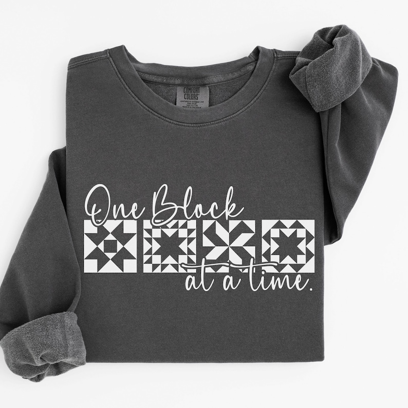 May include: Dark gray sweatshirt with the text "One Block at a Time" in white script and block letters. The design includes four white quilt block patterns. The sweatshirt has a crew neck and long sleeves.