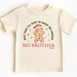 May include: Cream-colored toddler t-shirt with a gingerbread man graphic wearing a Santa hat. The text reads "Next year I'll have to share the cookies" and "Big Brother to be". The design includes snowflakes.