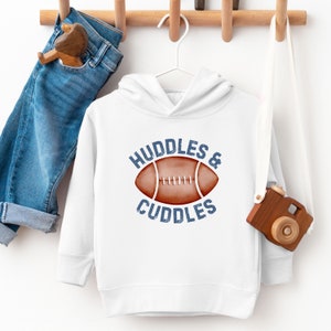 May include: White hoodie with the text "HUDDLES & CUDDLES" and a football graphic in brown and white. The hoodie is hanging on a white hanger. Blue jeans and a wooden toy are also visible.