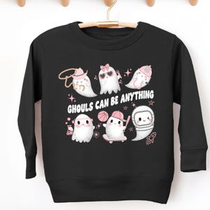May include: Black long-sleeved sweatshirt with cartoon ghosts in different outfits, such as a cowboy hat, sunglasses, and a space helmet. The white text reads "GHOULS CAN BE ANYTHING". The design is finished with pink stars and accents.