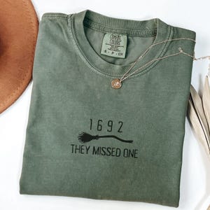 May include: An olive green t-shirt with the text "1692 THEY MISSED ONE" printed in black. A gold-colored necklace with a circular pendant rests on the shirt. The shirt is folded and placed on a white surface.