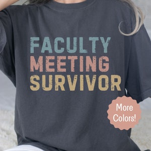 May include: A dark gray t-shirt with the text "FACULTY MEETING SURVIVOR" in a vintage style font with faded colors. The text is in a stacked format with "FACULTY" in blue, "MEETING" in pink, and "SURVIVOR" in yellow. The shirt has a distressed look and feel. The text "More Colors!" is in a white circle with a pink outline.