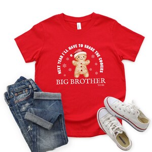 May include: A red t-shirt with the text "NEXT YEAR I'LL HAVE TO SHARE THE COOKIES" and "BIG BROTHER TO BE". The shirt features a gingerbread man wearing a Santa hat. The image also shows blue jeans and white sneakers.