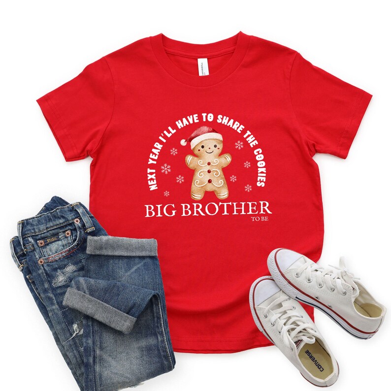May include: A red t-shirt with the text "NEXT YEAR I'LL HAVE TO SHARE THE COOKIES" and "BIG BROTHER TO BE". The shirt features a gingerbread man wearing a Santa hat. The image also shows blue jeans and white sneakers.