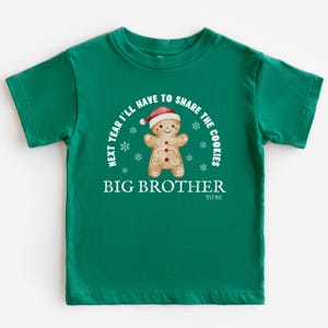 May include: A green toddler t-shirt with the text "NEXT YEAR I'LL HAVE TO SHARE THE COOKIES" and "BIG BROTHER TO BE". The shirt features a gingerbread man wearing a Santa hat and snowflakes.