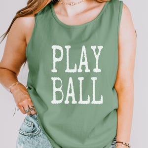 Puede incluir: Una camiseta sin mangas de color verde salvia con el texto PLAY BALL impreso en una fuente blanca desgastada en el centro del pecho. La prenda tiene un cuello redondo y un corte est&aacute;ndar, confeccionada en tela de color s&oacute;lido.
