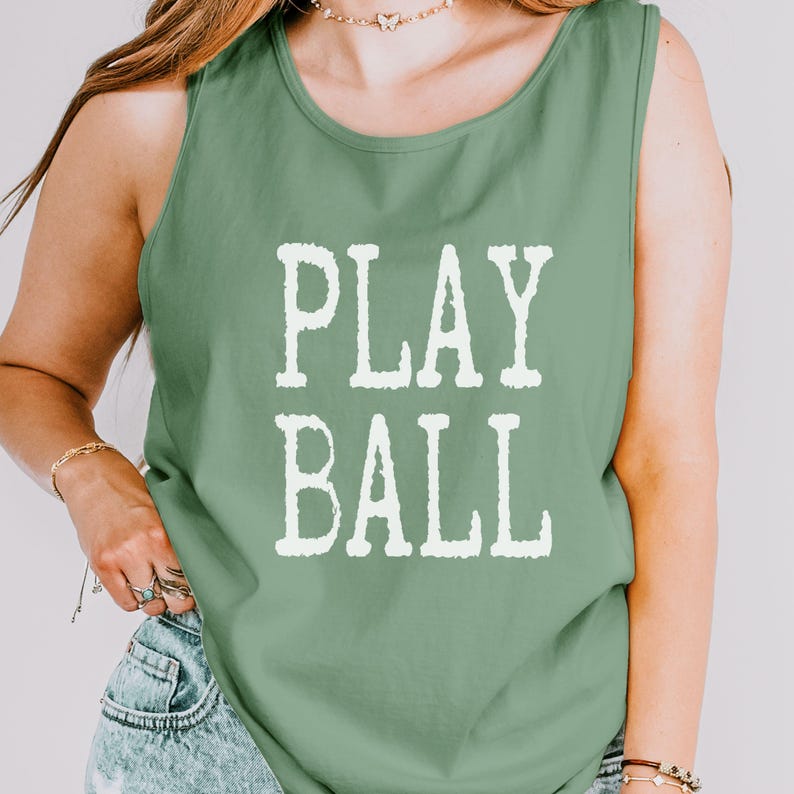 Puede incluir: Una camiseta sin mangas de color verde salvia con el texto PLAY BALL impreso en una fuente blanca desgastada en el centro del pecho. La prenda tiene un cuello redondo y un corte est&aacute;ndar, confeccionada en tela de color s&oacute;lido.
