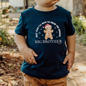 May include: Navy blue t-shirt with a gingerbread man graphic wearing a Santa hat. The text reads "Next year I'll have to share the cookies" and "Big Brother to be". The shirt is for a child.