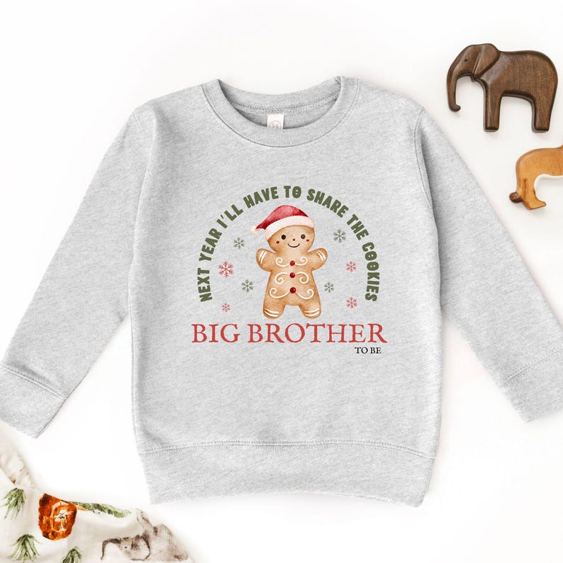 May include: A light gray sweatshirt with a gingerbread man graphic wearing a Santa hat. The text reads "Next year I'll have to share the cookies" and "Big Brother to be". Festive holiday apparel.
