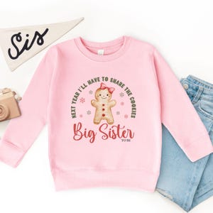 May include: A light pink sweatshirt with the text "Next year I'll have to share the cookies" and "Big Sister to be". The design includes a gingerbread person with a pink bow. A white pennant with "Sis" is in the background.