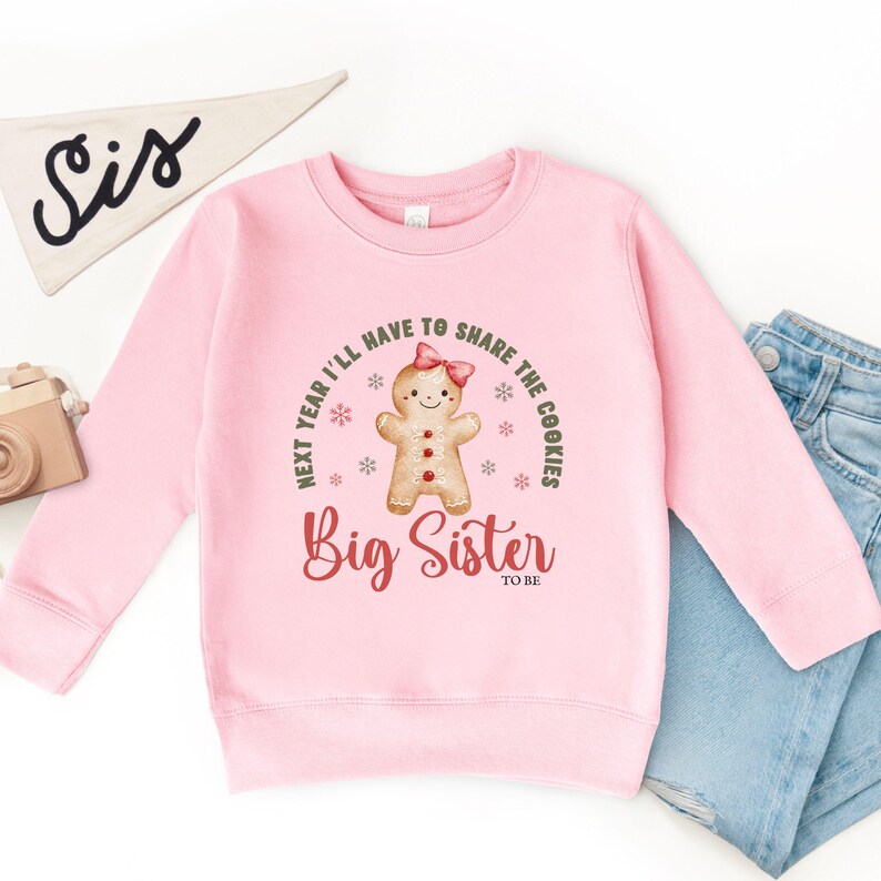 May include: A light pink sweatshirt with the text "Next year I'll have to share the cookies" and "Big Sister to be". The design includes a gingerbread person with a pink bow. A white pennant with "Sis" is in the background.