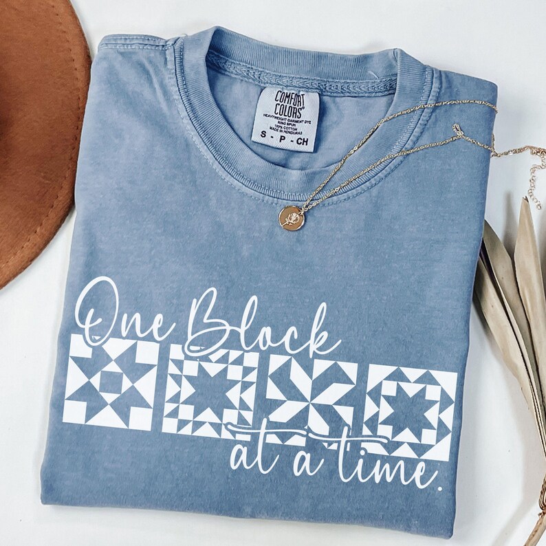 May include: A blue Comfort Colors t-shirt with the text "One Block at a time" and quilt block designs in white. A gold necklace with a pendant rests on the shirt. The shirt is folded and ready to wear.